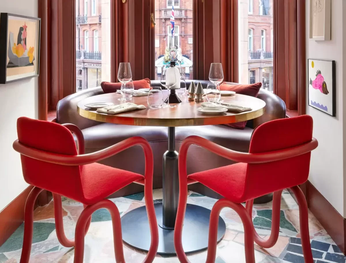 A cosy dining area features a round wooden table set with glassware and plates. Two red, wavy chairs are positioned on a patterned floor. The table is placed by large windows, with framed art and plush cushions completing the inviting ambiance at Mount St Restaurant, London.