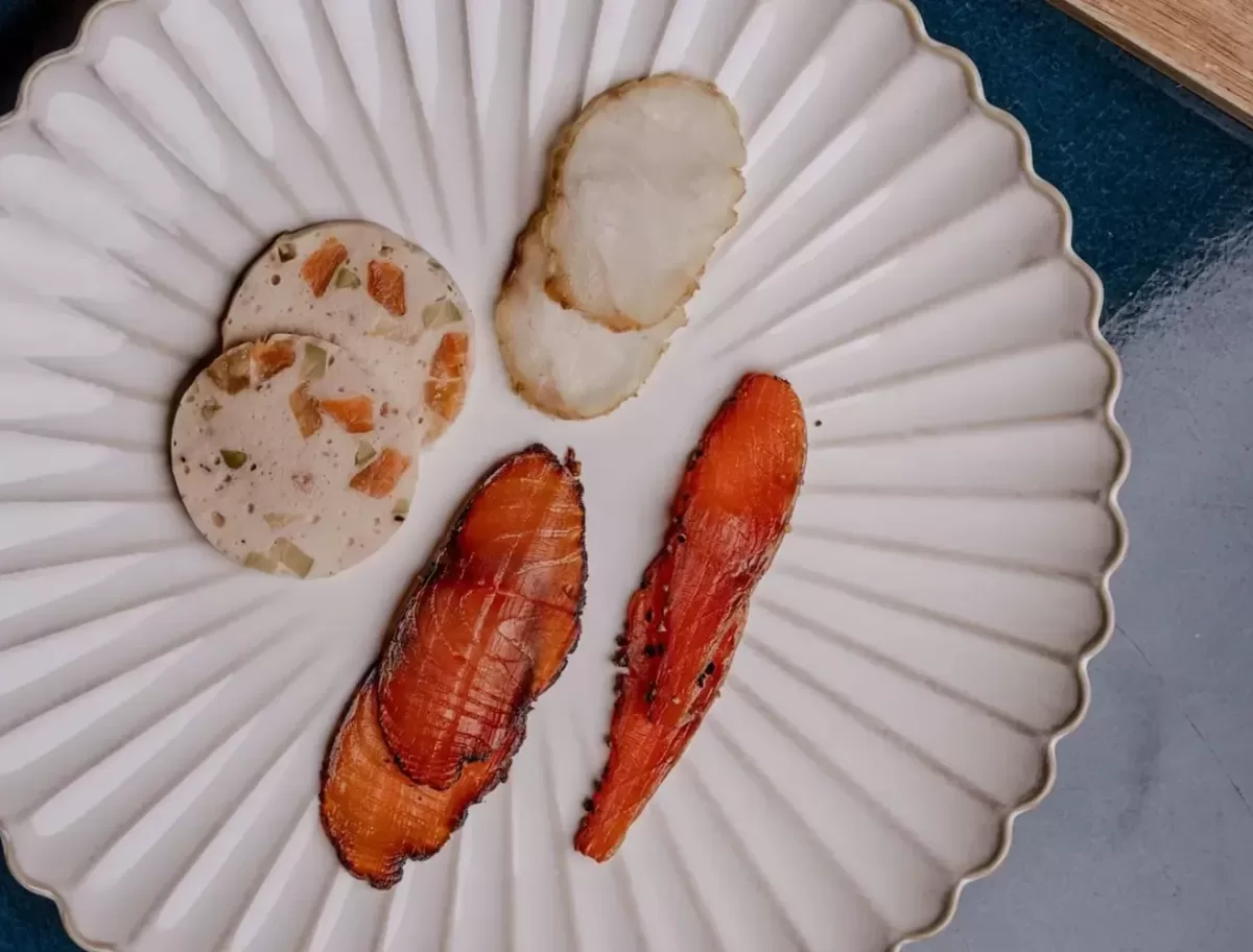 A white plate with a scalloped edge displays gourmet food: sliced terrine with visible vegetables, a piece of white fish, and two pieces of seared salmon. The plate rests on a dark blue surface.