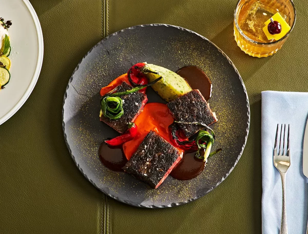 A dish with seared meat, drizzled with red and brown sauce, garnished with roasted red peppers and greens on a black plate. An avocado slice is at the top. Beside it, a glass with an orange liquid and ice, garnished with a cherry, is visible.