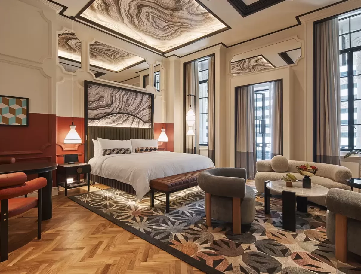 A luxurious hotel room with a large bed, geometric-patterned carpet, and modern furnishings. The ceiling features decorative panels, and the room is illuminated by stylish pendant lights and large windows.