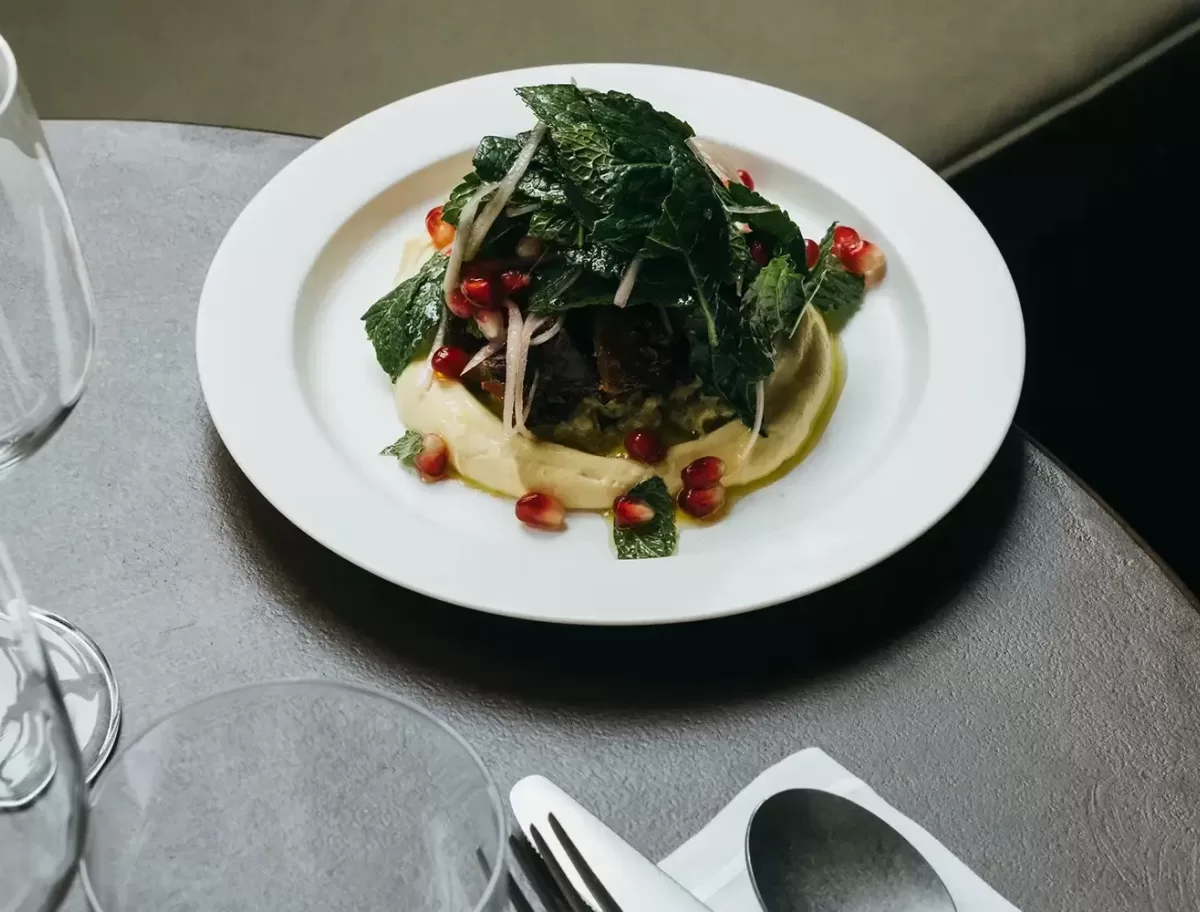 A dish plated on a white round plate, featuring meat topped with leafy greens and garnished with scattered pomegranate seeds. Its served with a light-colored puree. Wine glasses and a spoon are visible on the table beside it.