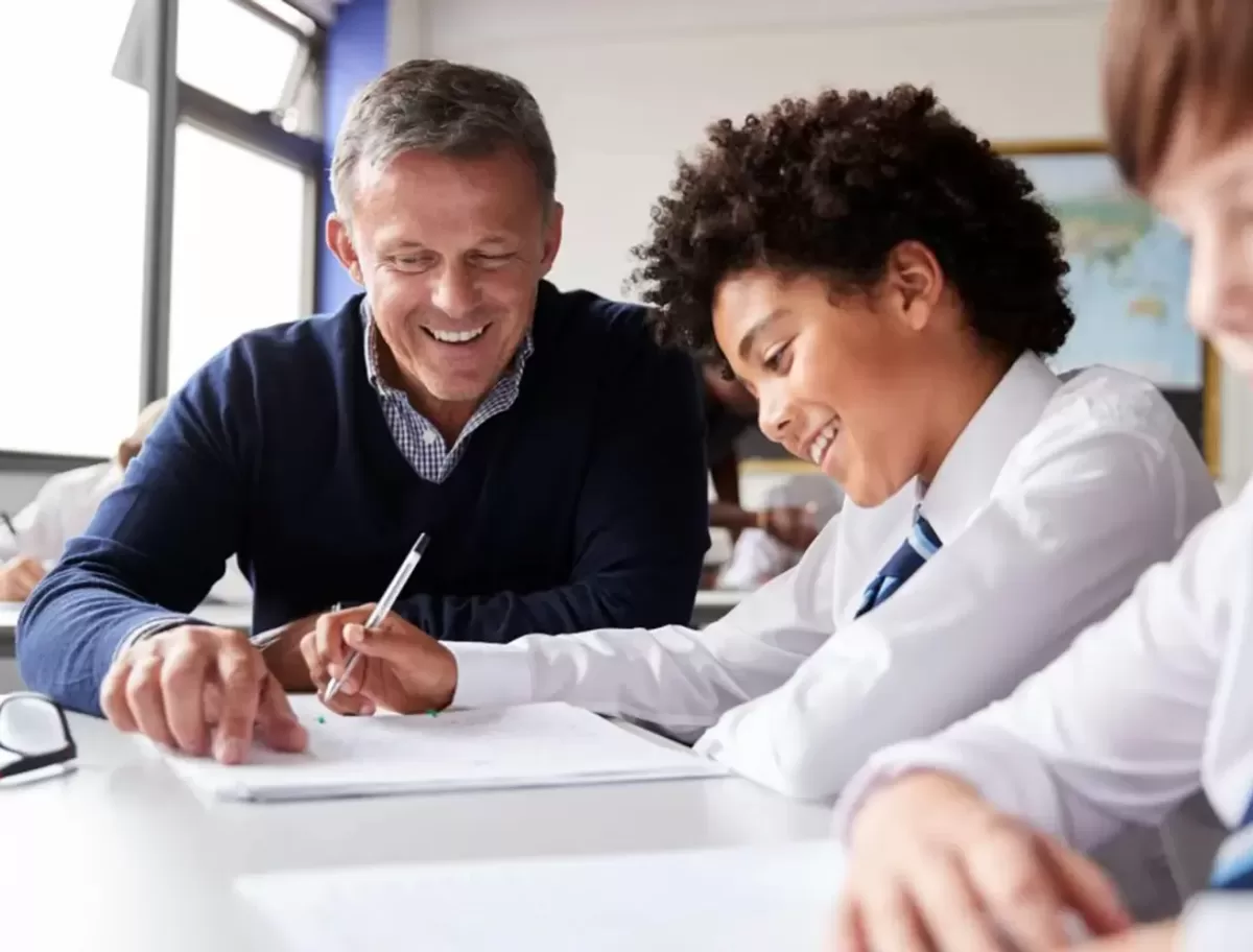Male teacher and students smiling, sitting at desk in a school completing work with a pen and paper.
