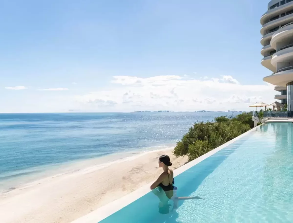 A person in a swimsuit sits at the edge of an infinity pool overlooking a serene beach and ocean. The sky is clear with a few clouds, and a modern building is visible to the right. The scene is peaceful and sunlit.