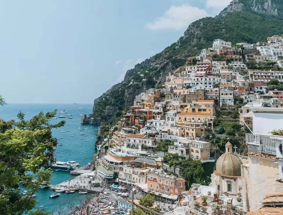 Colorful buildings stacked on a steep hillside overlooking a sparkling blue sea with boats, lush greenery, and a crowded beach below; mountains rise in the background under a clear, sunny sky.