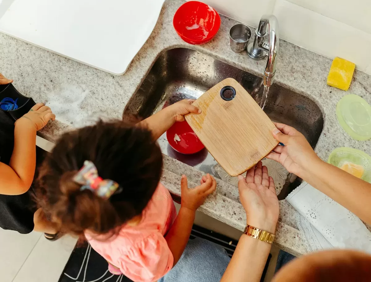 A child and an adult are washing a small wooden cutting board in a kitchen sink. The countertop is cluttered with soap, sponges, and a red bowl. The child is wearing a pink shirt, and the adult is wearing a watch on the left wrist.