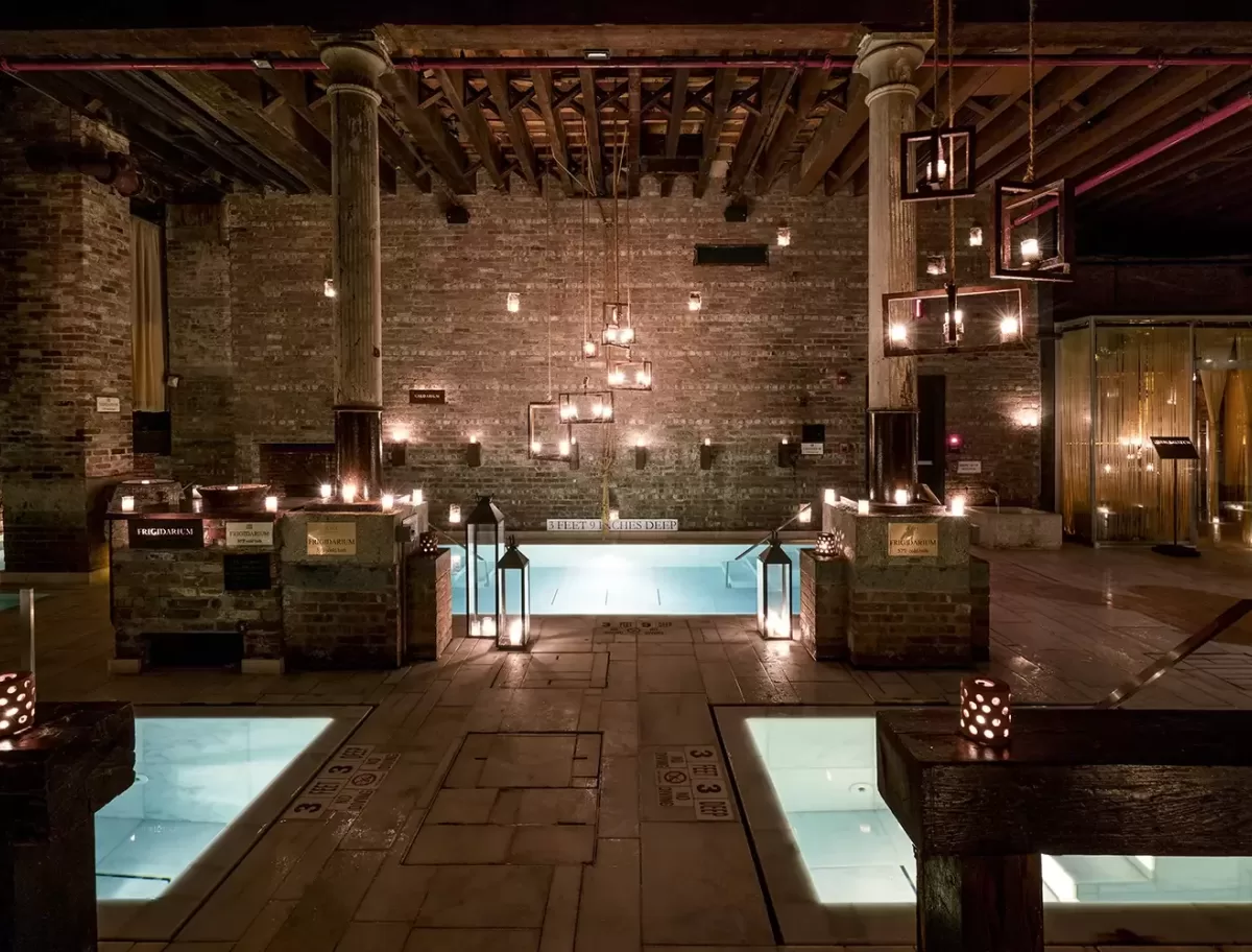 Spa interior with dim lighting features several rectangular pools surrounded by rustic stone walls and wooden beams. Candles and hanging lanterns illuminate the serene space. Steps lead into the central pool.