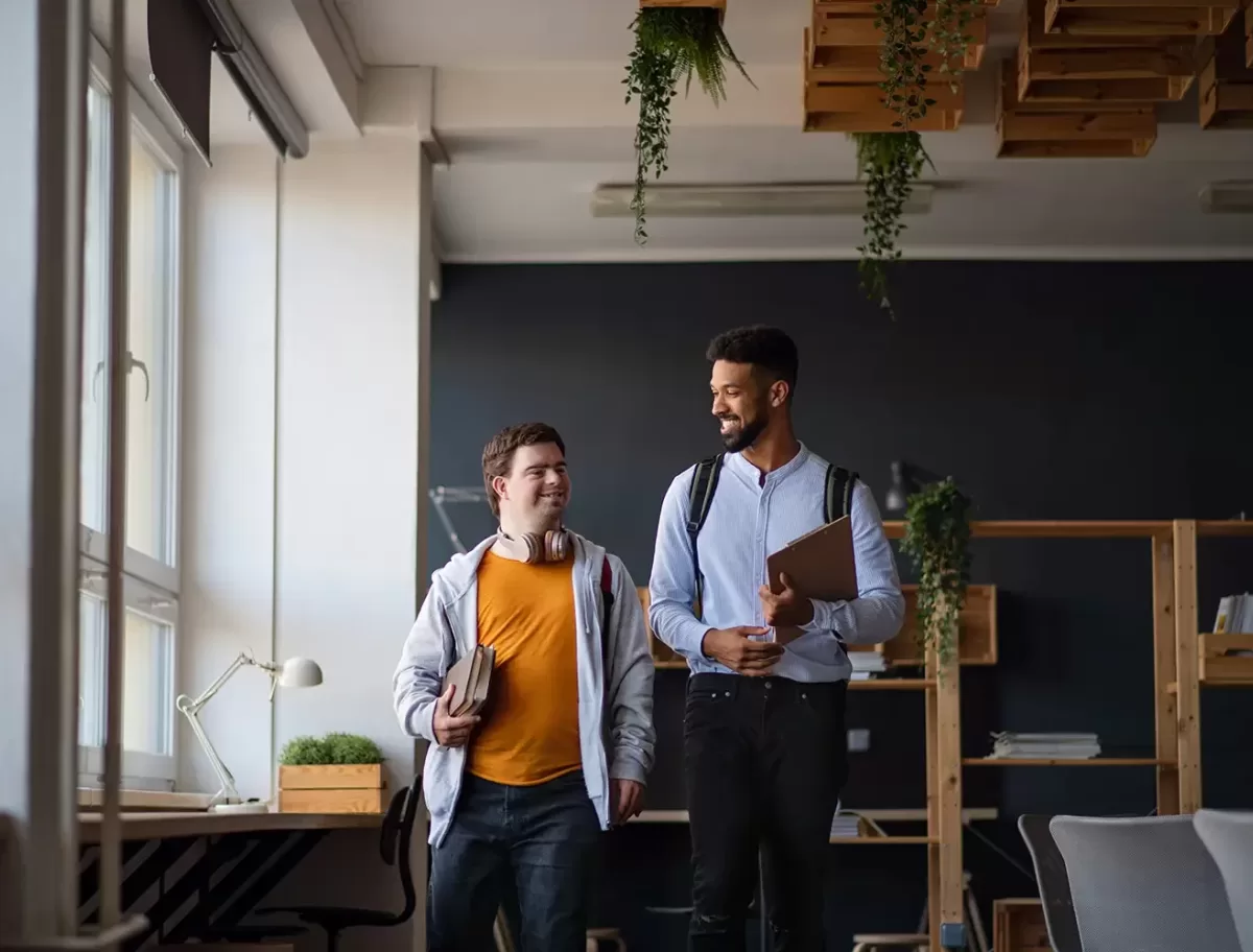 Two people walking and smiling in a modern office space with shelves and plants. One holds a laptop and a tablet, while the other carries books. Bright window light illuminates the room.