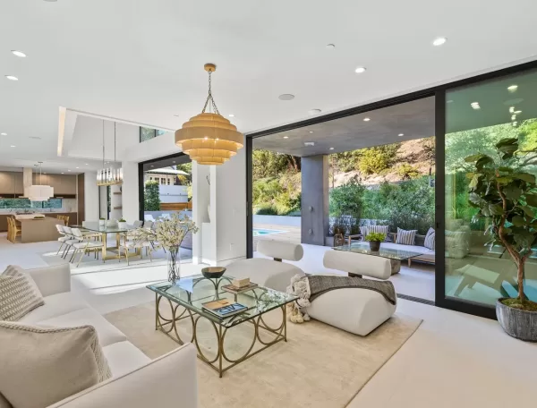 Modern open-concept living room with white furniture, glass coffee table, and gold chandelier. Large sliding doors open to a patio with seating, a pool, and lush greenery outside. Dining area and kitchen are visible in the background.