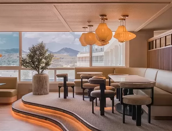 A modern, stylish lounge with tiered seating and a large potted plant. Pendant lights hang from the ceiling, and large windows offer a scenic view of mountains. Soft lighting creates a cozy ambiance.