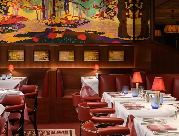 A cosy restaurant with red leather booths, white tablecloths, and red lamps on tables. The walls are adorned with colorful landscape art and framed paintings, creating a warm and inviting atmosphere.