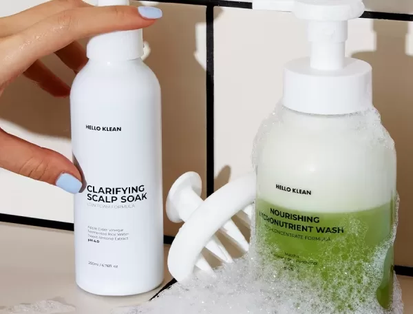 A hand holding a white bottle labeled Clarifying Scalp Soak next to a pump dispenser with green liquid labeled Nourishing Micro-Nutrient Wash. Both products are on a tiled surface with bubbles around them.