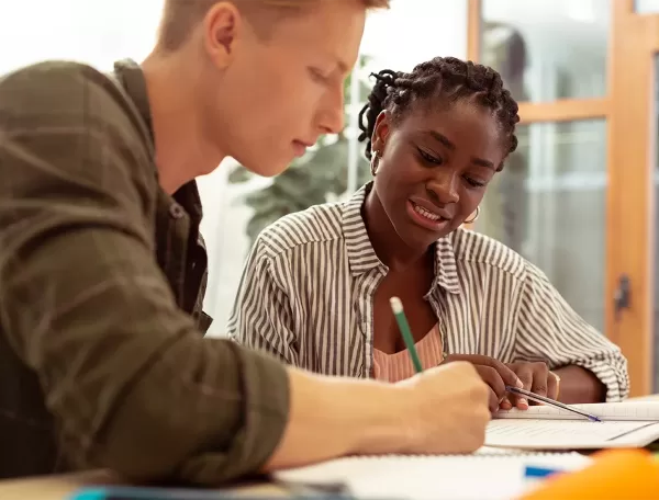 A young man and woman are sitting at a table, working together on notebooks. The man is writing, while the woman smiles and looks on. They appear to be in a study or collaborative session, with books and stationery around them.