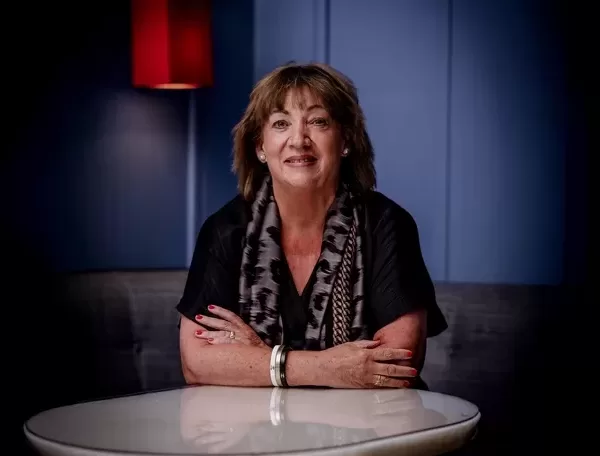 Photo of Paula Fitzherbert (Maybourne’s global head of communications) with shoulder-length brown hair and a patterned scarf sitting at a round table, arms crossed. She is smiling warmly against a backdrop of a blue wall and a red lamp.
