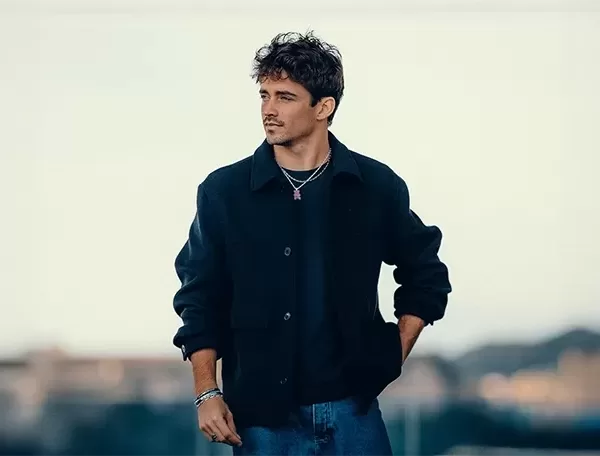 Charles Leclerc - with curly hair stands outdoors wearing a dark jacket, black shirt, and jeans. He has a thoughtful expression, with his right hand in his pocket. The background is blurred, showing a soft, neutral sky and some indistinct structures.