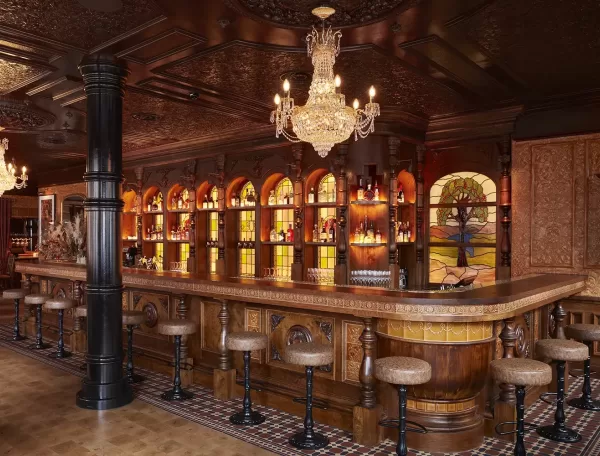 A luxurious, vintage-style bar with a wooden counter, padded barstools, a crystal chandelier, stained glass artwork, and shelves of bottles illuminated by warm lighting. Ornate patterns decorate the ceiling and walls.