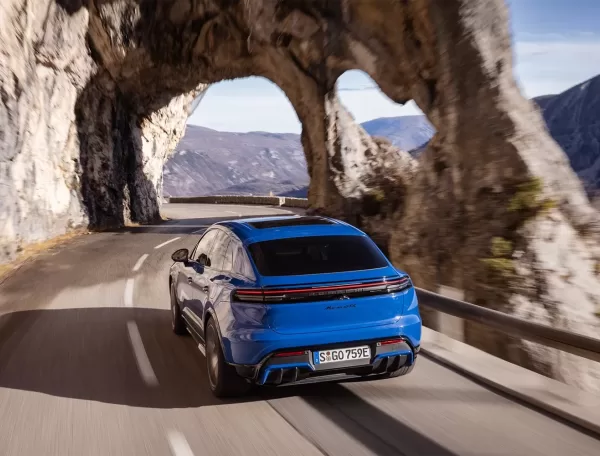 A blue sports car drives along a winding mountain road, passing through a natural rock arch with scenic cliffs and distant hills in the background under a partly cloudy sky.