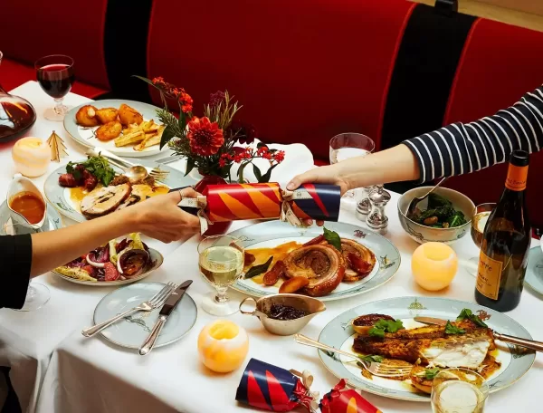 A festive dinner table with two people pulling a Christmas cracker. Plates feature roasted meat, vegetables, and a side dish. The table is decorated with candles, flowers, and a glass of white wine at Ham Yard Hotel, Soho in London.