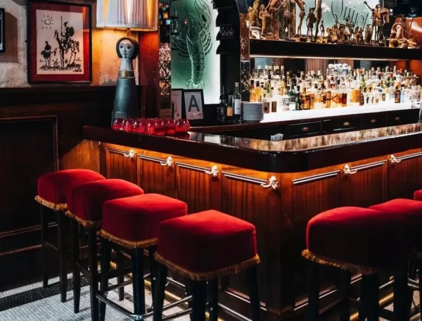 A cosy bar in NYC with a polished wooden counter and red-cushioned barstools. The shelves behind are stocked with various bottles, and decorative art pieces adorn the walls. The ambiance is warm and inviting.