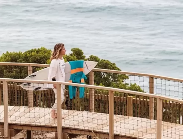 Women stood on a wooden walkway bridge across beach next to the sea holding a surf board at Ericeira, Portugal