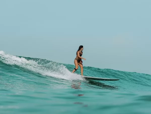 A woman in a swimsuit is surfing on a wave in the ocean. She is standing on a surfboard, riding along a crest with water splashing around. The sky is clear, adding to the serene and adventurous atmosphere.