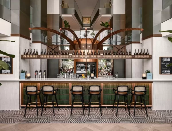 Modern bar interior with a long counter lined by six chic bar stools. Shelves stocked with bottles are displayed above, flanked by two chalkboards. The flooring has a geometric pattern, and large plants decorate the space.