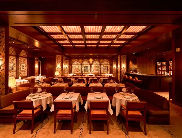 Elegant restaurant interior with warm lighting, featuring rows of tables set with white tablecloths and wooden chairs. The decor includes patterned wallpaper, framed artwork, and an inviting bar area in the background.