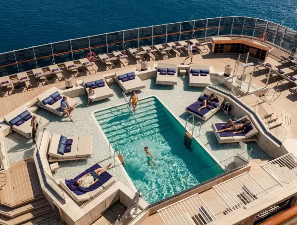 A view from above of a cruise ship’s pool deck, with people relaxing on lounge chairs and swimming in a small pool surrounded by clear railings and the ocean in the background.