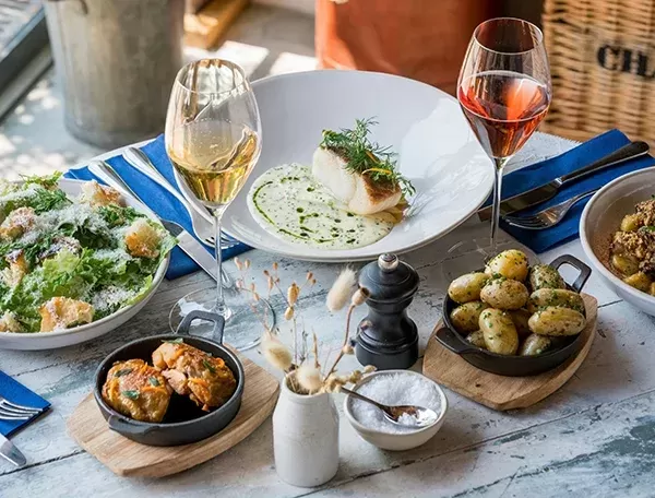 A delicious spread of dishes on a rustic table, featuring a salad, seared fish with sauce, roasted potatoes, and chicken stew, accompanied by two glasses of wine. A pepper mill and small vase add a cozy touch.