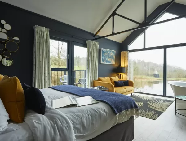 A modern bedroom with large windows overlooks a scenic outdoor landscape. The room features a bed with a blue blanket, a yellow sofa, circular mirrors on the wall, and a comfortable chair. Natural light fills the space, creating a cosy atmosphere.