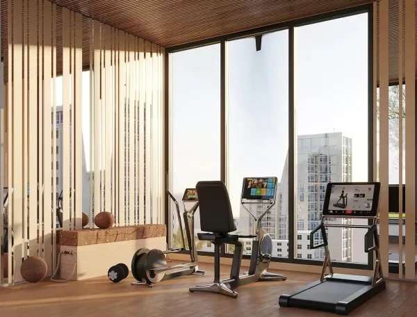A modern gym room with large windows overlooking tall buildings, featuring a treadmill, two stationary bikes, exercise balls, and wooden decor elements on a wood floor.