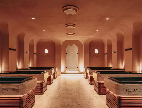 A warmly lit, modern spa room with ten marble soaking tubs arranged in two rows, arched ceilings, and soft ambient lighting creating a calm, inviting atmosphere. The word REBASE is displayed on the tubs and walls.