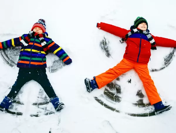 Two children dressed in colorful winter clothes lie on their backs making snow angels. The child on the left wears a striped jacket, and the one on the right wears an orange and red outfit. Both smile joyfully against the snowy backdrop.