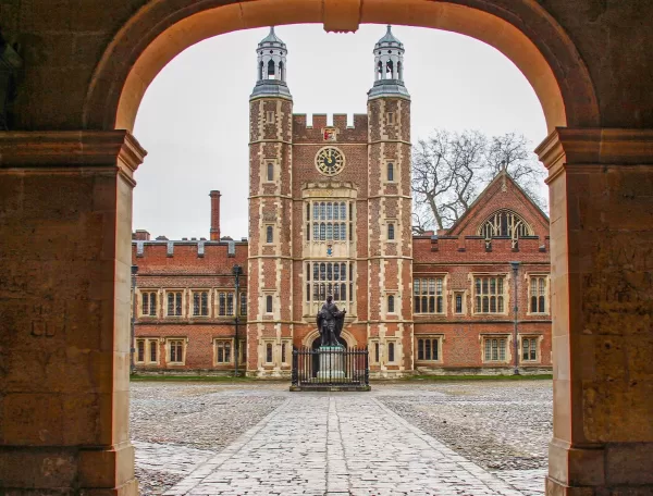 View through an archway of a historic brick building with a clock tower, central statue, cobblestone courtyard, and gothic architectural details on a cloudy day.
