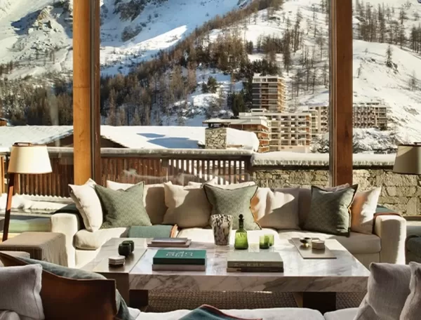 Cozy living room with a large wooden-framed window offering a scenic view of snow-covered mountains and trees. The room features comfortable beige sofas adorned with cushions around a central coffee table with decorative items.