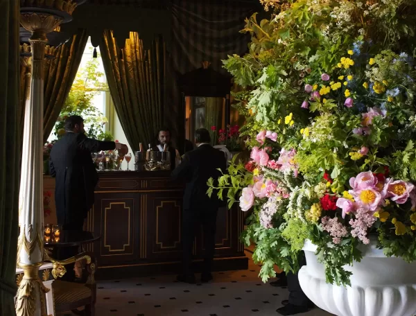 Three people in suits stand at a dark wood bar in an elegant room with heavy drapes. A large, vibrant flower arrangement in a white vase dominates the foreground on the right. Natural light comes in through a window.