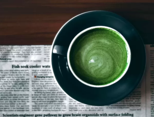 A cup of green matcha latte seen from above, placed on a dark saucer and sitting on a newspaper on a wooden table. The latte forms a swirling pattern in the middle of the cup.