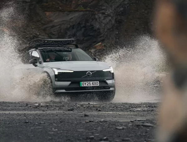 A white SUV with a roof rack drives through a large puddle, splashing water on a rocky, rugged road with mountains in the background. The cars headlights are on and the license plate reads OY25 NKO.