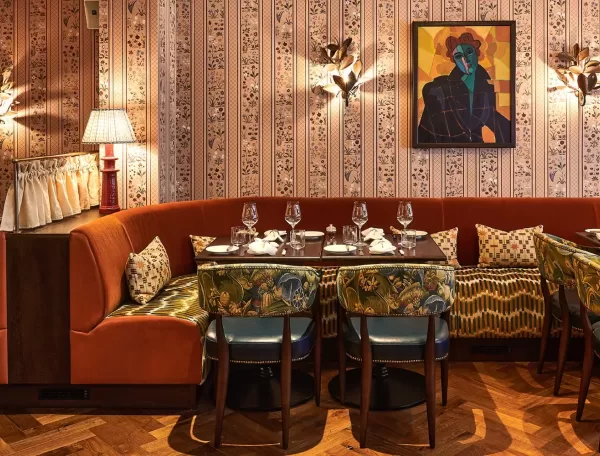 Cosy restaurant interior featuring a curved orange booth with patterned pillows, surrounded by green, floral-upholstered chairs, a patterned wallpaper, and a colorful abstract painting. Tables are set with glasses and napkins.