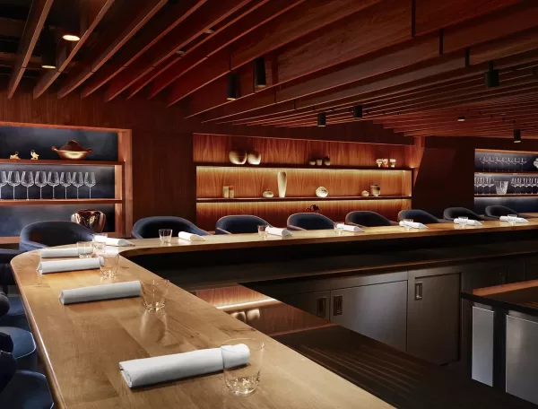 The modern, elegant restaurant interior of Kitchen Table, Fitzrovia, with a long, wooden counter surrounded by navy blue chairs. Soft, warm lighting highlights glassware and decorative items on wooden shelves, creating a cozy, sophisticated ambiance.
