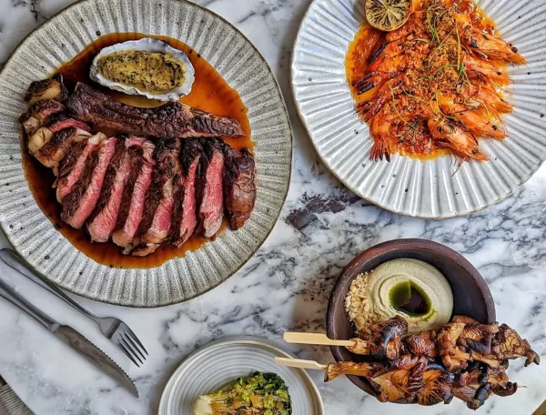 A marble table with plates of food: sliced steak with sauce, grilled prawns with lemon garnish, skewered grilled chicken with hummus, and a small bowl of greens. Silverware is placed beside the plates at Milk Beach, London.