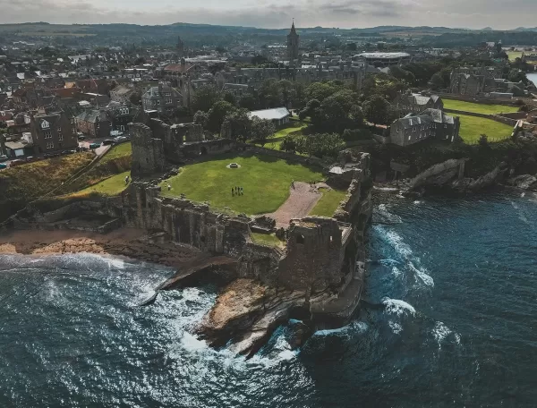 Aerial view of coastal ruins of a large stone castle, surrounded by green lawns, beside the sea. A historic town with many stone buildings stretches out behind the castle under a cloudy sky.