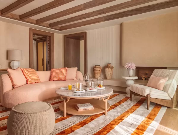 A cosy living room with a pink sofa and orange-checkered pillows. Theres a round coffee table with drinks and snacks, a striped armchair, and decorative vases. The room features wooden beams and a patterned rug in warm tones in Temuda Bay.