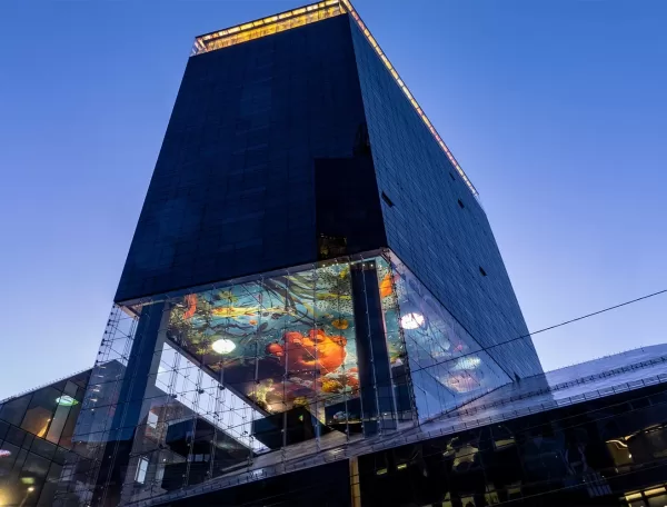 A modern, tall building with a reflective glass facade is shown against a clear blue sky. A vibrant digital display underneath a protruding section features colorful abstract art, visible from the street below.