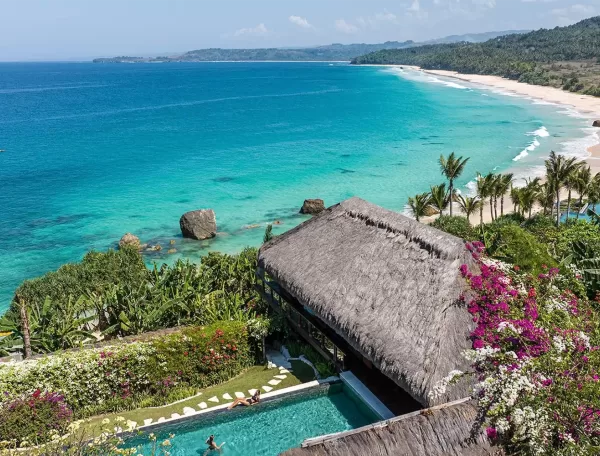 A scenic view of a tropical coastline with turquoise waters and a sandy beach. In the foreground, a thatched-roof structure and a pool surrounded by vibrant flowers and greenery. Hills are visible in the distance under a clear blue sky.