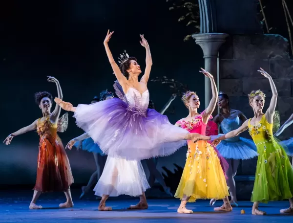 A ballerina in a purple and white tutu stands on stage, surrounded by dancers in tutus. They hold sparkling wands, poised elegantly on a staircase backdrop, creating a magical, fairytale-like scene at Royal Ballet & Opera’s Cinderella.