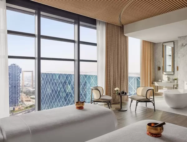 A beautiful room with floor to ceiling windows overlooks Dubai. Inside the room is 2 white massage beds with a golden sound bowl placed on each bed.