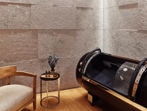 A minimalist room featuring a wooden chair with a beige cushion, a small round table with artful decorations, and a sleek black pod-like structure, all set against textured stone walls at Longevity at Bvlgari Spa.