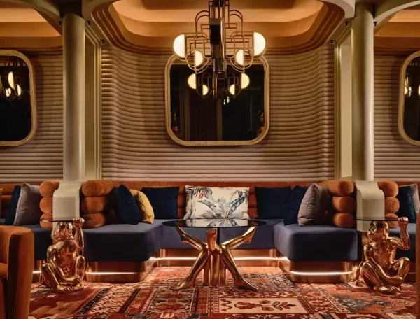 Stylish lounge with a luxurious interior featuring a navy blue sectional sofa, ornate chandelier, geometric wall patterns, a glass coffee table with a star-shaped gold base, and gold dog sculptures. Richly patterned rug covers the floor.