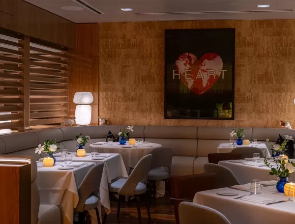 A cozy, dimly lit restaurant with white tablecloths, modern chairs, floral centerpieces, and glowing candles. A large heart artwork with the word HEART hangs on the wall behind a curved booth.