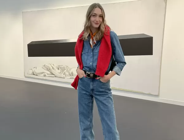 A woman stands indoors wearing a denim shirt and jeans, with a red sweater draped over her shoulders. She poses with one hand in her pocket, smiling slightly. Behind her is a large, minimalist artwork on the wall.